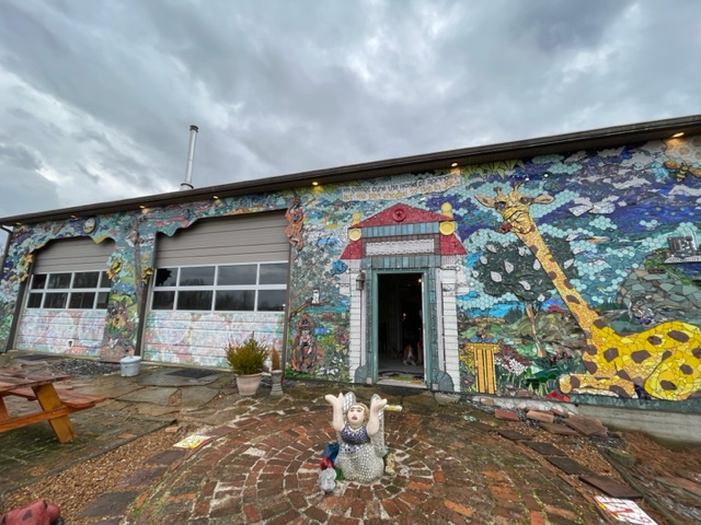 This image shows the facade of the Bell Buckle Mosaic House with a focus on a kneeling angel sculpture named JOY on the front patio.