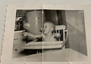 An image of an old black and white Polaroid photo of a small child, Lynn, in a highchair holding a large flower with a look of awe and excitement.