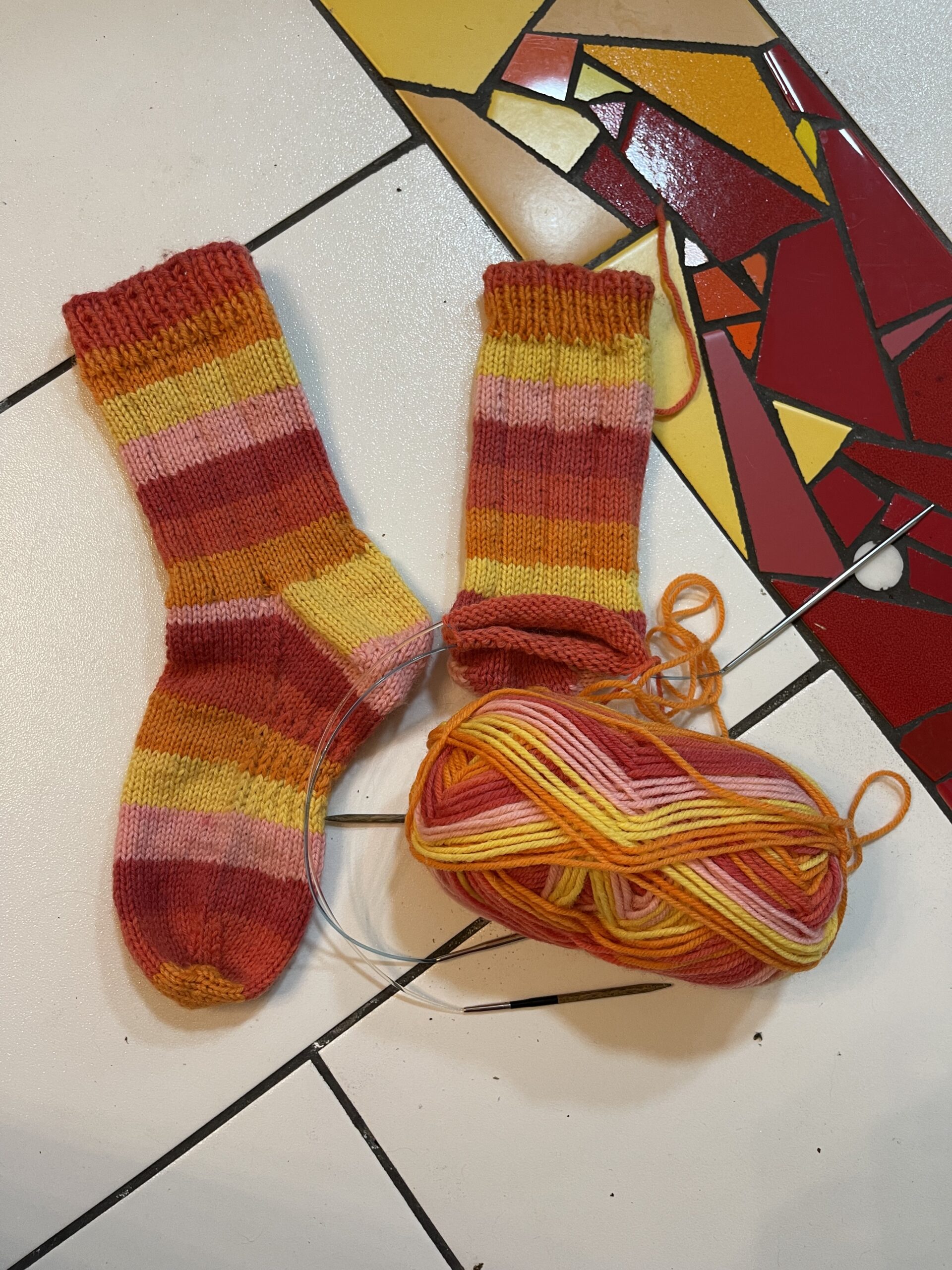 The image is of a pair of red, orange, yellow and pink striped knit socks with one completed and the other still on the needles.