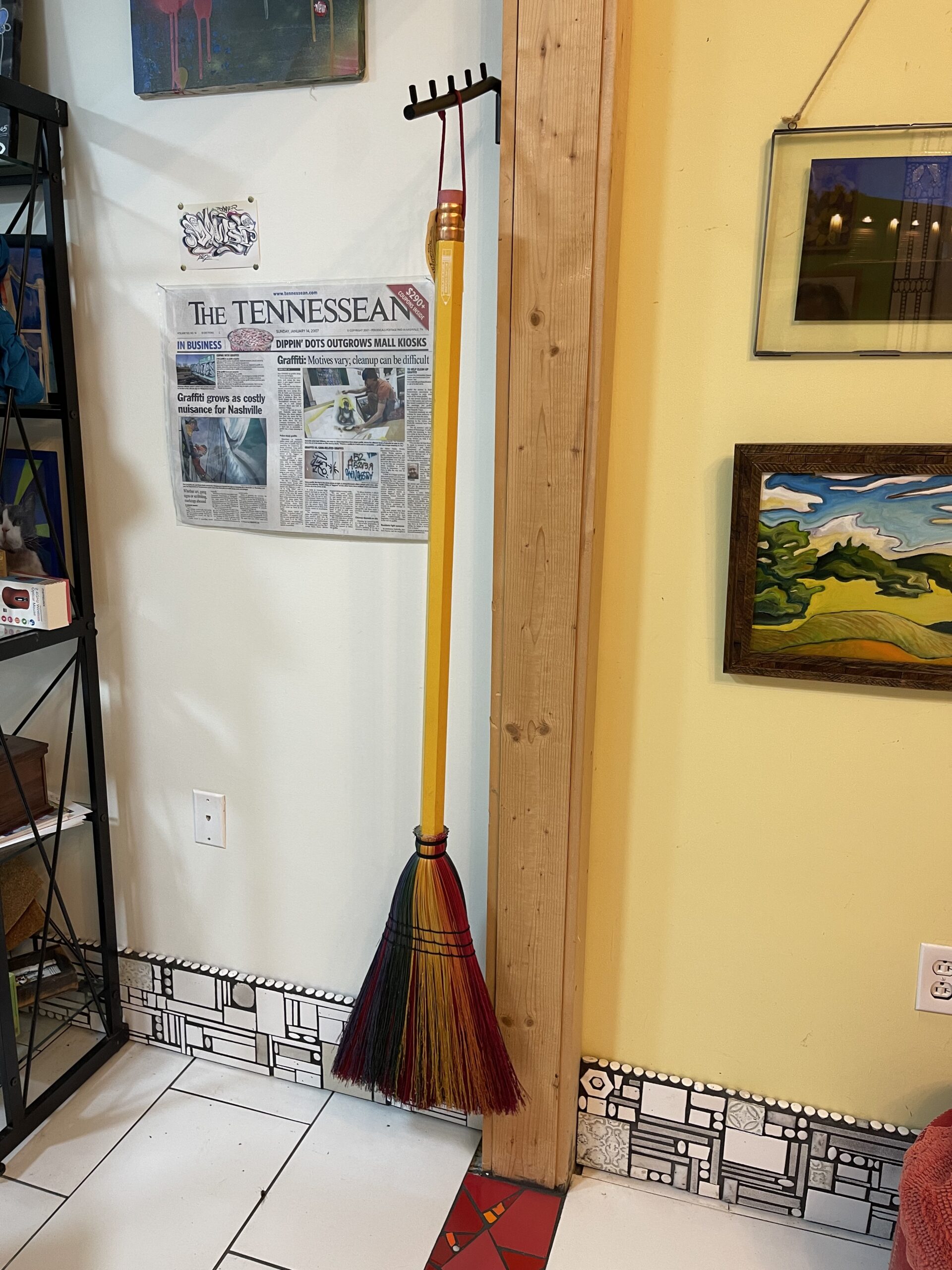 The image is a broom hanging on a peg. The handle of the broom is a large pencil and the bristles are rainbow colors.